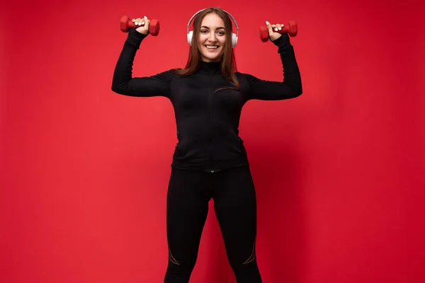Beautiful happy smiling young brunet woman wearing black sport clothes isolated on red background doing fitness using dumbbells wearing white bluetooth headsets listening to music looking at camera — Stock Photo, Image