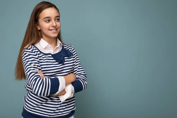 Güzel, pozitif gülümseyen esmer, çizgili uzun kollu bir kız çocuğu. Mavi arka plan duvarının üzerinde tek başına el ele tutuşup yan tarafa bakıyor. — Stok fotoğraf