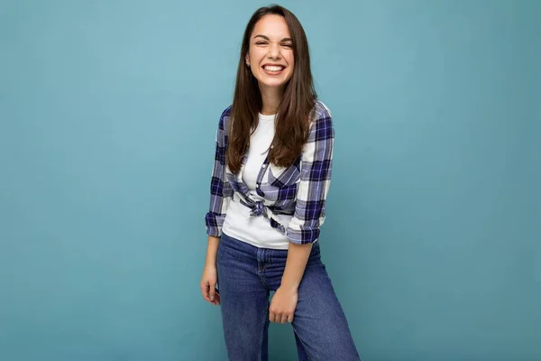 Portrait photo de jeune belle femme brune hipster souriante en chemise et jeans bleu et blanc tendance. Sexy insouciante femme posant isolé près du mur bleu avec espace vide en studio — Photo