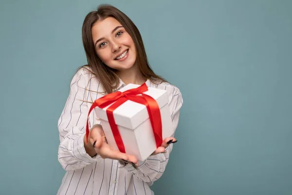 Tiro de atractiva mujer rubia oscura sonriente positiva aislada sobre la pared de fondo colorido que usa ropa de moda cotidiana sosteniendo la caja de regalo y mirando a la cámara —  Fotos de Stock