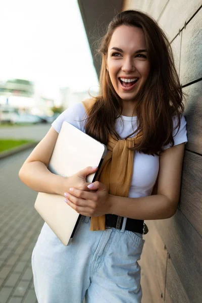 Krásný šťastný brunet mladá žena hledá na straně v ulici s notebookem v rukou s bílým tričkem a modré džíny — Stock fotografie