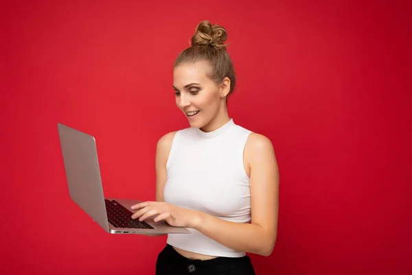 Side profile photo shot of beautiful smiling happy blond young woman with gathered hair wearing white t-shirt using computer laptop typing on keyboard looking at netbook monitor screen isolated over — Stock Photo, Image