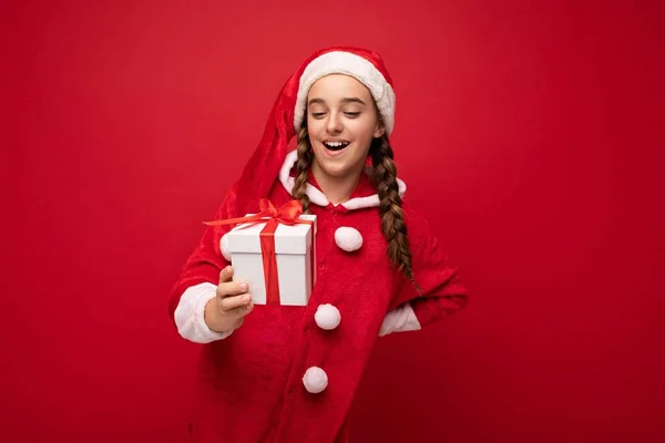 Skott av attraktiva glada leende brunett flicka isolerad över röd bakgrund vägg bär Santa Claus kostym håller vit presentförpackning med rött band och tittar på överraskning — Stockfoto