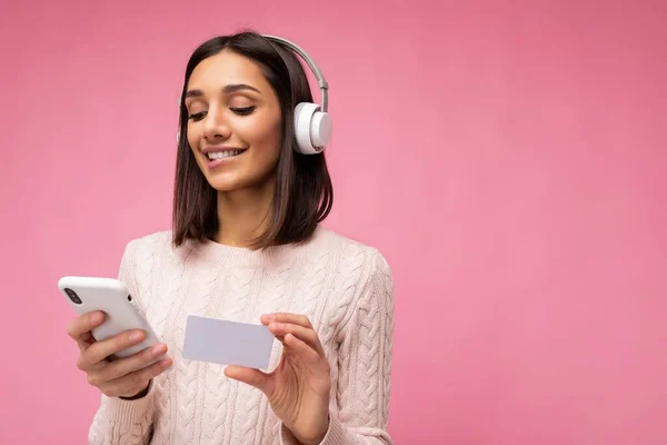 Photo of beautiful positive smiling young brunette woman wearing pink casual sweater isolated over pink background wall wearing white bluetooth wireless headphones and listening to music and using — Stock Photo, Image