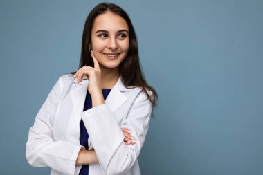 Çekici, güzel, mutlu, düşünceli, gülümseyen esmer kadının portresi. Samimi duyguları olan, mavi arka planda izole edilmiş beyaz bir sağlık önlüğü giyen, fotokopi çeken.