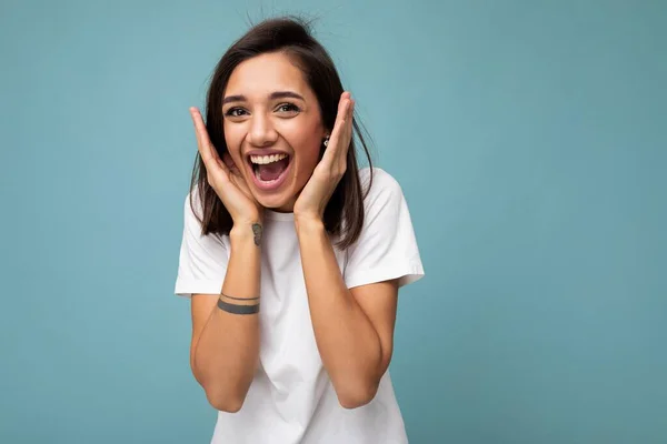 Porträtt av glada positiva leende förtjusande ung vacker brunett kvinna med uppriktiga känslor bär casual vit t-shirt för mockup isolerad på blå bakgrund med kopia utrymme — Stockfoto