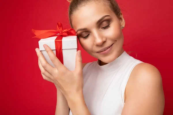 Portré fénykép vonzó fiatal szőke nő elszigetelt felett piros háttér fal visel fehér felső gazdaság ajándék doboz és élvezze csukott szemmel — Stock Fotó
