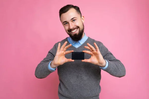 Bonito sorridente morena barbuda homem vestindo camisola cinza e camisa azul isolado na parede de fundo segurando cartão de crédito olhando para a câmera — Fotografia de Stock