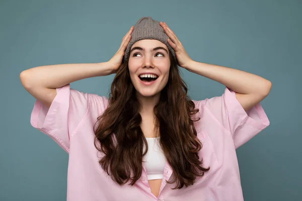 Foto der schönen emotionalen jungen brünetten Frau, die isoliert auf blauem Hintergrund Wand trägt rosa Hemd und grauen Hut, der zur Seite schaut — Stockfoto