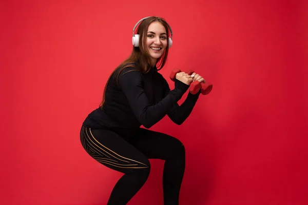 Side-profile photo of beautiful positive smiling young brunette woman wearing black sport clothes isolated on red background wall squating using dumbbells wearing white bluetooth earphones listening — Stock Photo, Image