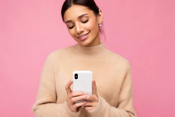 Szekrény portré Vonzó aranyos mosolygós fiatal barna nő visel bézs meleg pulóver áll elszigetelt felett rózsaszín háttér szörfözés az interneten telefonon keresztül nézi a mobil képernyőn — Stock Fotó