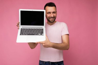 Laptop bilgisayarı tutan yakışıklı adam pembe arka plandaki tişörtlü kameraya bakıyor.