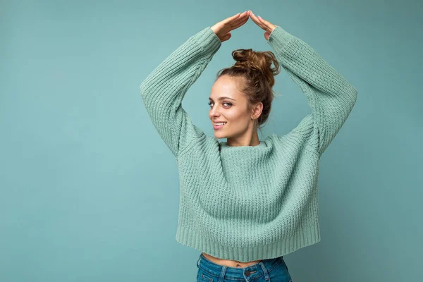 Portrait of young happy positive beautiful blonde woman with sincere emotions wearing casual blue pullover isolated on blue background with copy space and showing house gesture with hands — Stock Photo, Image
