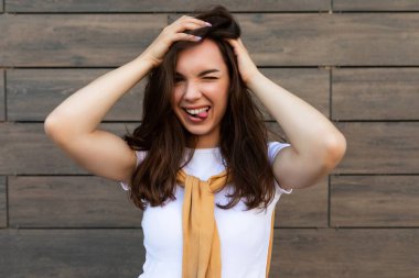 Başarılı gülümseyen mutlu esmer kadının portresi. Günlük beyaz tişört ve kot pantolon giyiyor. Sokakta kahverengi duvarın yanında zehirleniyor ve eğleniyor.