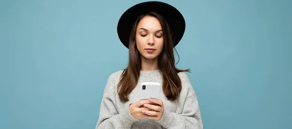 年轻美丽的黑发女子头戴黑色帽子，头戴灰色毛衣，手持智能手机，低头看手机发短信，背景孤立 — 图库照片