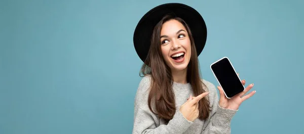 Panoramic Closeup of charming young happy woman wearing black hat and grey sweater holding phone looking to the side pointing finger at screen isolated on background — Stock Photo, Image