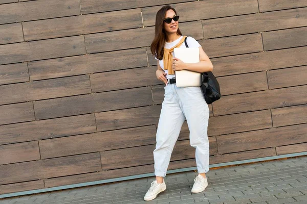 Full length photo shot of young charming beautifyl brunet woman going in the street near brown wall looking to side in white t-shirt and light blue jeans white sneakers with black female bag holding — Stock Photo, Image