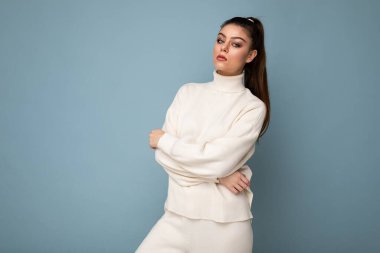 Beyaz renkli süveter giyen güzel, genç, esmer bir kadın, elleri önünde katlanmış, göğsüne yakın, mavi arka planda, kopya alanı ile izole edilmiş.