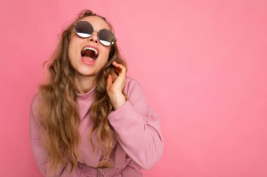 Pembe arka plan duvarında izole edilmiş güzel, aşırı neşeli, esmer, kıvırcık saçlı bir kadının fotoğrafı. Günlük pembe spor kıyafetleri ve şık güneş gözlükleriyle yukarı bakıyor ve eğleniyor.