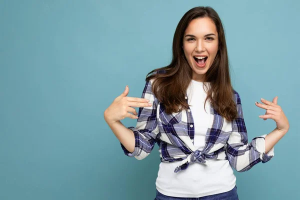 Billeder af ung positiv sjov glædelig smuk brunette kvinde med oprigtige følelser iført trendy check shirt stående isoleret på blå baggrund med ledig plads - Stock-foto