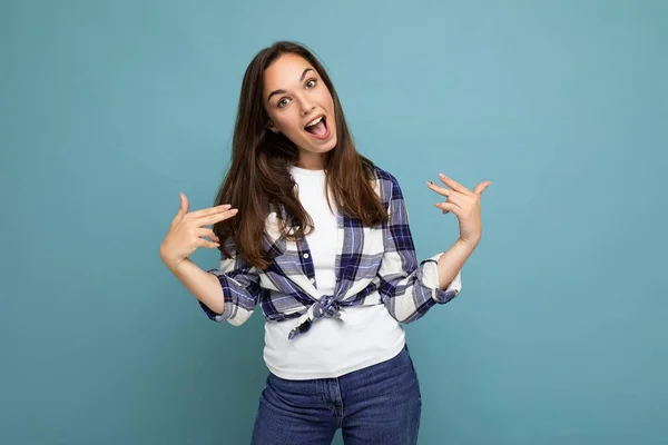 Shot van jonge positieve heerlijke glimlachende mooie brunette vrouw met oprechte emoties dragen trendy check shirt staan geïsoleerd op blauwe achtergrond met vrije ruimte en wijzen naar jezelf — Stockfoto