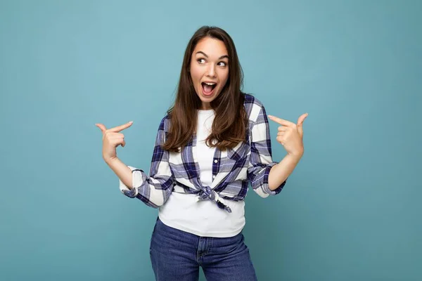 Jonge positieve heerlijke aantrekkelijke brunette vrouw met oprechte emoties dragen trendy check shirt geïsoleerd op blauwe achtergrond met vrije ruimte en wijzend naar jezelf — Stockfoto