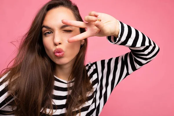 Photo of young positive happy beautiful brunette woman with sincere emotions wearing casual striped pullover isolated on pink background with copy space and showing peace gesture and giving kiss — Stock Photo, Image
