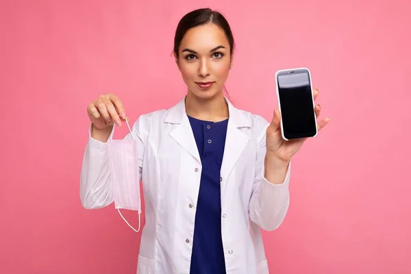 Beyaz önlüklü çekici genç bir kadın pembe arka planda izole edilmiş modern cep telefonunu kameraya bakarak gösteriyor. — Stok fotoğraf