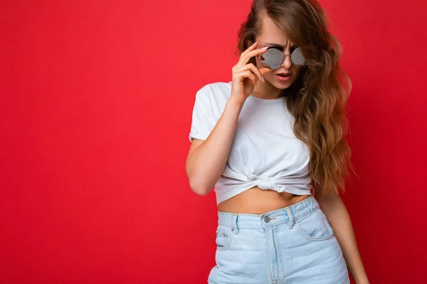 Tiro de encantador sexy jovem loira escura encaracolado mulher isolada sobre a parede de fundo vermelho vestindo casual t-shirt branca e óculos de sol elegantes olhando para a câmera — Fotografia de Stock