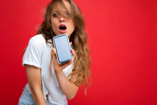 Foto von sexy überrascht junge blonde Frau gut aussehend trägt weißes T-Shirt isoliert auf rotem Hintergrund mit Kopierraum hält Telefon zeigt Smartphone in der Hand mit leerem Bildschirm für — Stockfoto