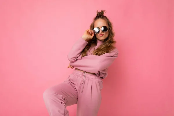Foto av glad positiv cool ung vacker blond vågigt hår kvinna med uppriktiga känslor bär snygga rosa sportkläder och solglasögon isolerad på rosa bakgrund med kopiera utrymme och ha kul — Stockfoto