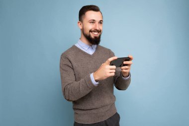 Arka plandaki duvarda izole edilmiş gri kazak ve mavi gömlek giyen, sakallı, mutlu, genç, esmer bir adamın cep telefonuyla oyun oynaması ve eğlenmesi.