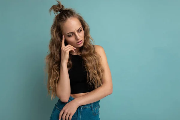 Photo of young thoughtful beautiful blonde curly woman with sincere emotions wearing black top isolated over blue background with copy space and thinking — Stock Photo, Image