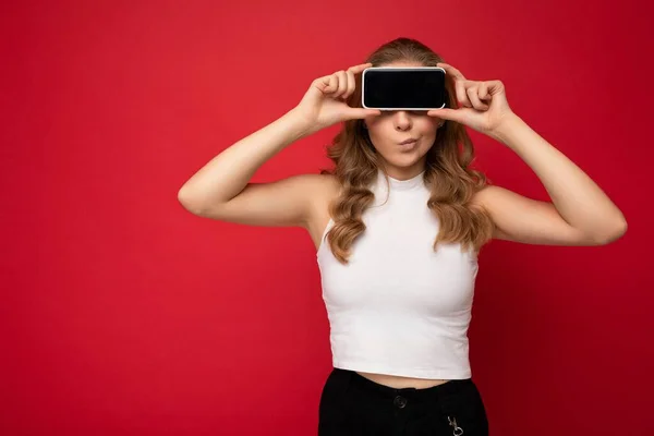 Engraçada jovem loira vestindo camiseta branca isolada no fundo vermelho com espaço de cópia segurando smartphone mostrando telefone na mão com tela vazia para recorte — Fotografia de Stock