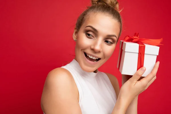Portré fotó vonzó boldog csodálkozó fiatal szőke nő elszigetelt felett piros háttér fal visel fehér felső gazdaság ajándék doboz és keres az oldalán — Stock Fotó