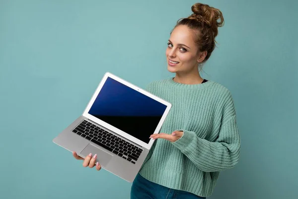 Frumoasă blondă întunecată tânără femeie cu păr creț adunat uitându-se la camera care deține laptop-ul computerului cu ecran de monitor gol cu batjocură în sus și spațiu de copiere purtând mânecă lungă albastră izolată pe albastru deschis — Fotografie, imagine de stoc