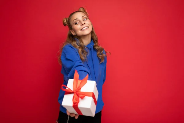 Fascinante feliz sorrindo incrível jovem loira isolada sobre a parede de fundo vermelho vestindo azul elegante capuz segurando caixa de presente e olhando para a câmera — Fotografia de Stock