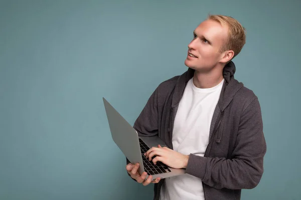 Boczny profil zdjęcie przystojnego blondyna trzymającego laptopa na klawiaturze myślącego o czymś w białym t-shircie i szarym swetrze patrzącego na netbook odizolowany na niebiesko — Zdjęcie stockowe