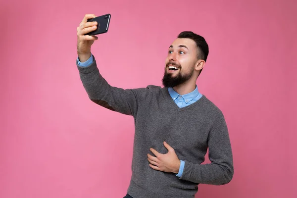 Фото красивой молодой брюнетки с небритым мужчиной с бородой в обычном сером свитере и синей рубашке, изолированной на розовом фоне, держащей смартфон с онлайн видео-звонком, смотрящим на — стоковое фото