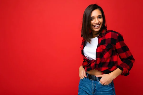 Portrét pozitivní veselý úsměv mladé brunetky žena v ležérní bílé tričko, stylové červené kontrolní tričko izolované na červeném pozadí s kopírovacím prostorem — Stock fotografie