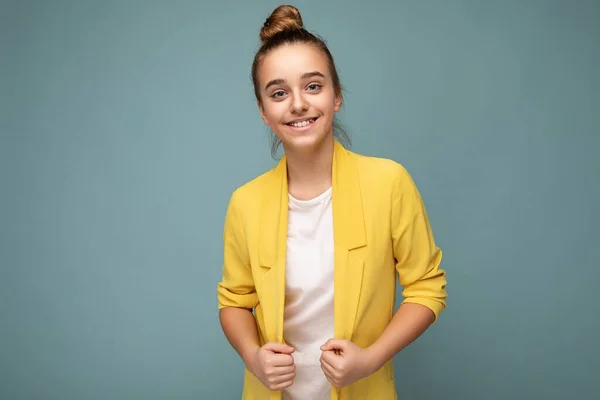 Снимок красивой счастливой улыбающейся брюнетки маленькая девочка в модной желтой куртке и белой футболке, стоящей изолированно над синей стеной, смотрящей в камеру — стоковое фото