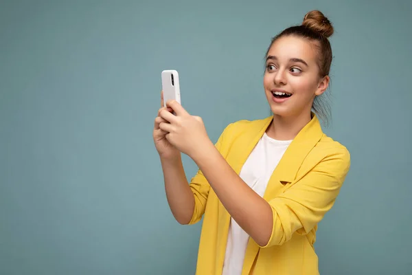 Hermosa joven sorprendida chica con chaqueta amarilla y camiseta blanca de pie aislado sobre fondo azul navegar en Internet a través del teléfono mirando a la pantalla móvil — Foto de Stock