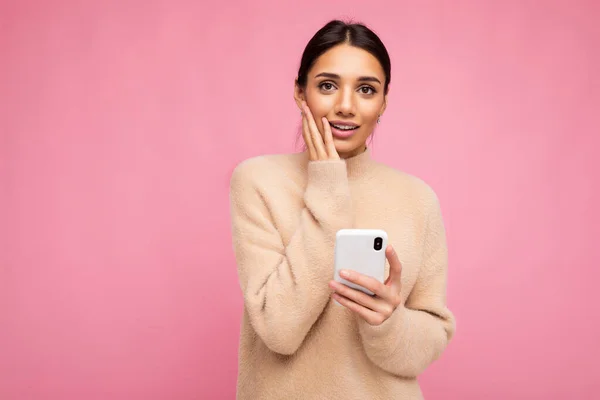 Vonzó aranyos szomorú fiatal barna nő visel bézs meleg pulóver áll elszigetelt felett rózsaszín háttér szörfözés az interneten telefonon keresztül nézi a kamera és az érintett — Stock Fotó