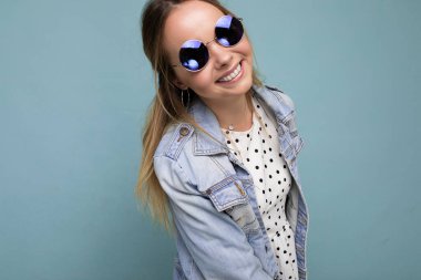 Çekici gülümseyen, mutlu, genç, sarışın kadın günlük şık kıyafetler giyiyor ve renkli arka plan duvarında izole edilmiş modern güneş gözlüğü takıyor.