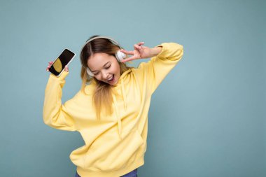 Çekici, pozitif gülümseyen, şık günlük kıyafetler giyen, renkli arka plan duvarında izole edilmiş, cep telefonlu beyaz bluetooth giyimli, boş ekranlı genç bir kadın.