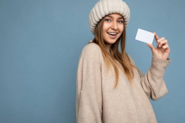 Güzel, mutlu, pozitif gülümseyen, koyu renkli, bej süveter ve örgü bej şapka giyen bir kadın. Mavi arka planda izole edilmiş ve kredi kartını kameraya bakarken gösteriyor.
