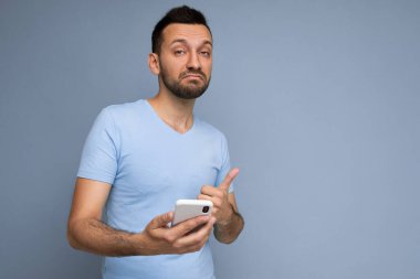 Düşünceli yakışıklı genç adam arka plan duvarından izole edilmiş günlük giysiler giyiyor ve cep telefonu yazıları yazıyor kameraya bakıyor ve düşünüyor.