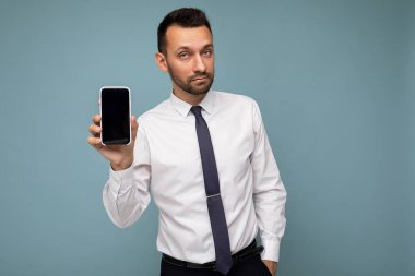 Yakışıklı, yakışıklı, yakışıklı, şık kıyafetler giyen, renkli arka plan duvarında tek başına duran, akıllı telefon tutan ve boş ekran ekranlı telefonu gösteren yakışıklı bir adam.