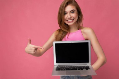 Güzel sarışın bir kadın elinde bilgisayar bilgisayarı olan boş bir monitör ile netbook 'a bakıyor pembe ekin bluzlu pembe arka planda izole edilmiş kameraya bakıyor.