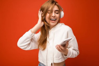 Güzel, neşeli, güler yüzlü, şık kıyafetler giyen, arka plan duvarından izole edilmiş, cep telefonu kullanan, beyaz bluetooth kulaklık takan ve müzik dinleyen genç bir kadının fotoğrafı.
